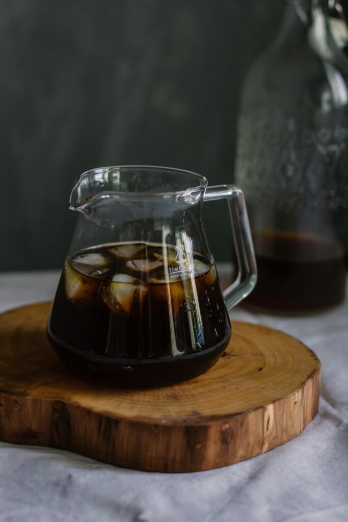 A glass pitcher filled with iced coffee on a round wooden board, creating a refreshing still life setup.