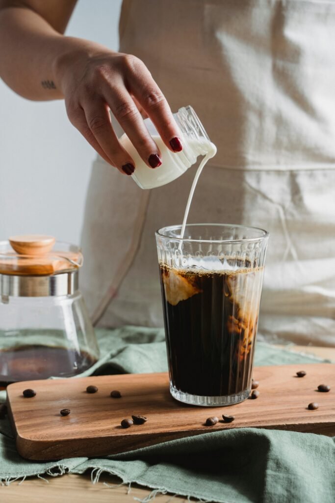 A person pours milk into a glass of cold brew coffee, surrounded by coffee beans, creating a captivating swirl.