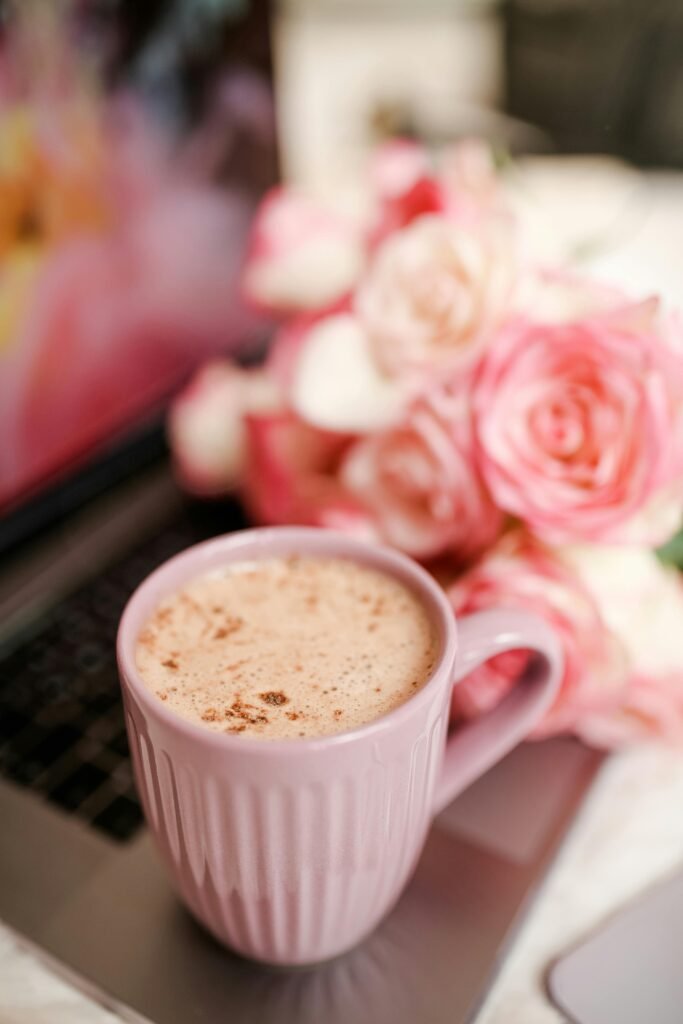 A close-up of a creamy latte in a pink cup with soft pink roses on a laptop.