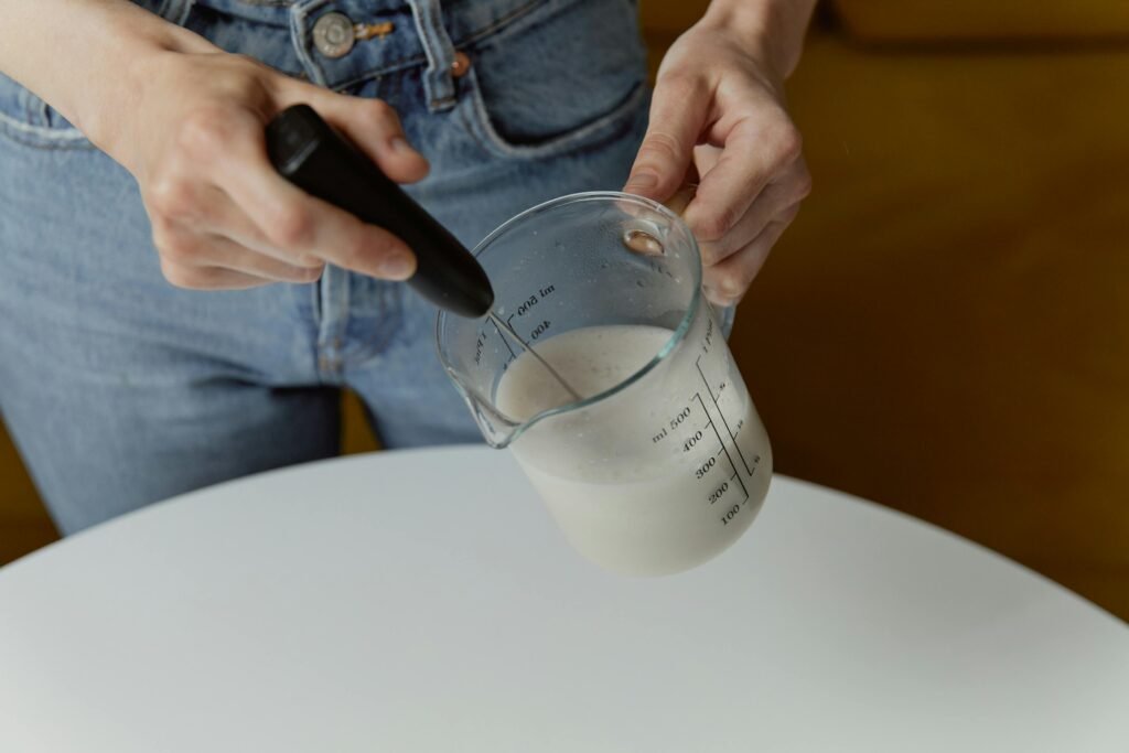 A person frothing milk with a handheld whisk in a transparent measuring cup, showcasing food preparation.