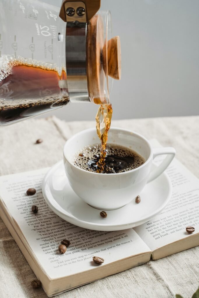 A steaming cup of coffee being poured from a pot over an open book, creating a cozy atmosphere.