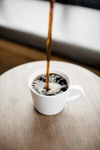 Steaming black coffee being poured into a white ceramic mug on a wooden table.