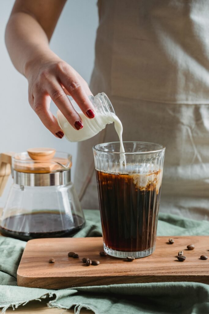 Close-up of a woman pouring milk into a cold brew coffee on a wooden board.