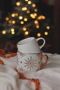 Decorative mugs with Christmas designs set against warm bokeh lights and festive decor.